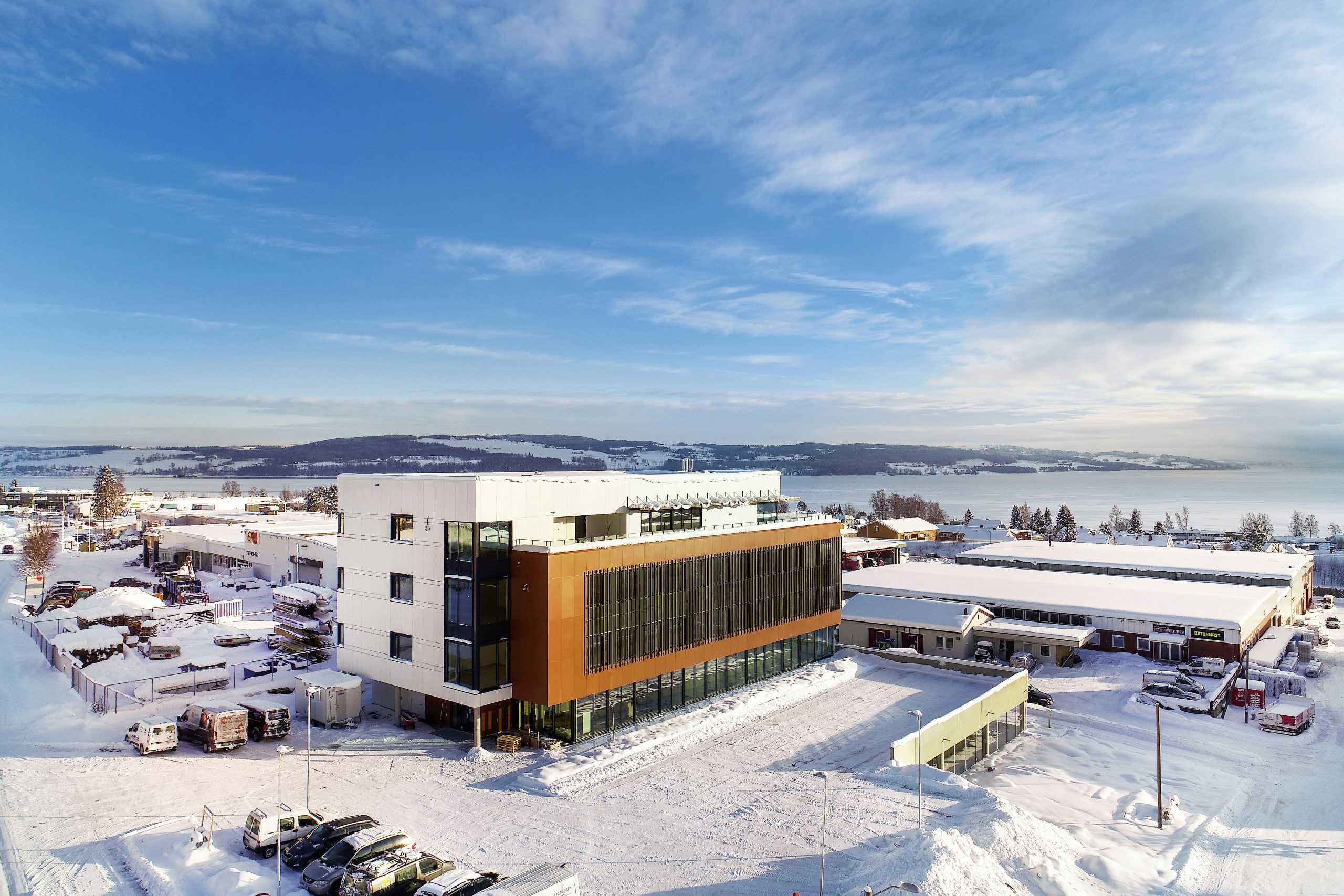 Foto av kontorbygg med en hvit del og en mørkere del, vinterlandskap med Mjøsa i bakgrunnen