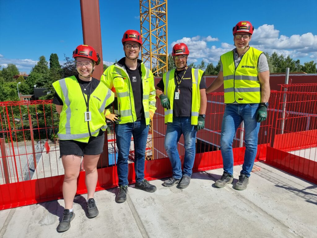 En kvinne, 3 menni synlighetstøy på byggeplass, blå himmel, byggekraner i bakgrunnen